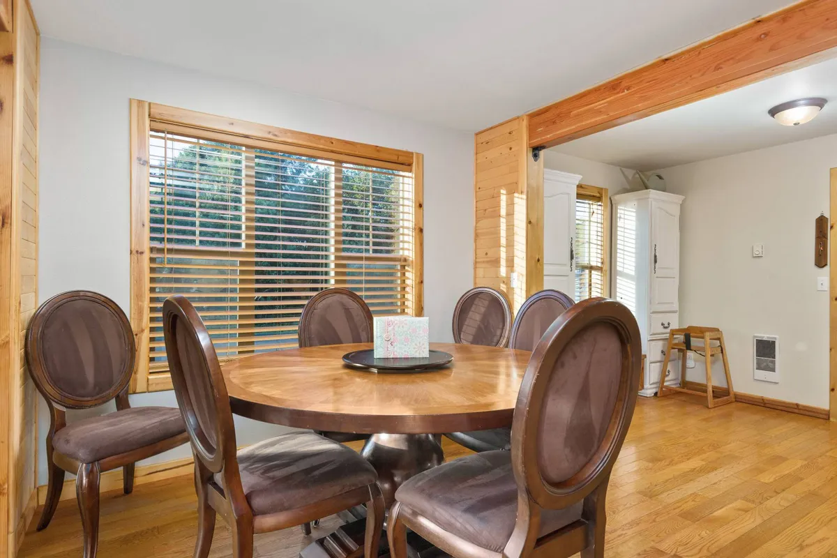 A round wooden dining table surrounded by upholstered chairs sits next to large windows with blinds, offering a comfortable space for family meals, celebrations, or quiet coffee mornings.