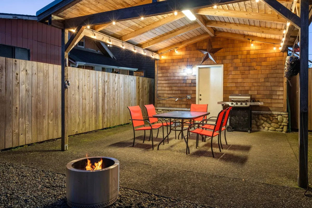 Another view of the backyard, showcasing an outdoor dining table with bold red chairs, a fire pit, a grill, and a wooden structure that provides shelter and ambiance. The scene emphasizes the spaciousness and functionality for gatherings and pet play.