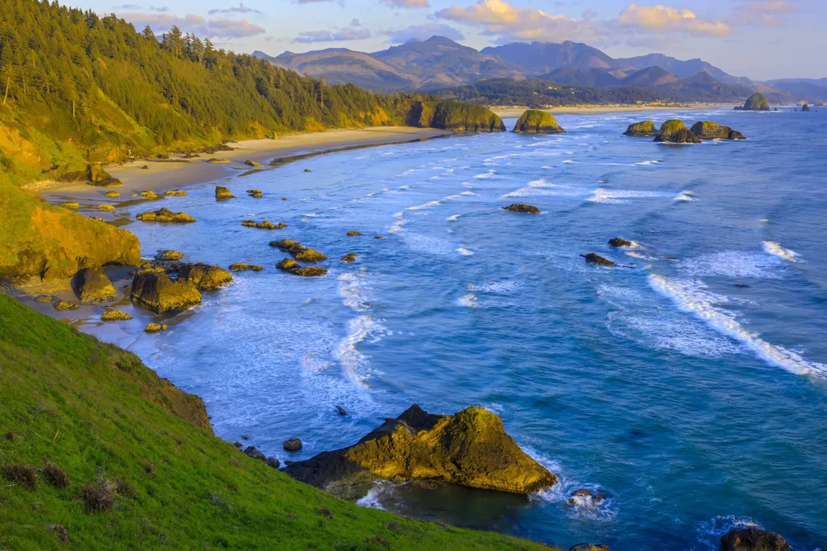 A serene coastal view featuring gentle ocean waves crashing on the shore, unique rock formations, and distant mountains. The tranquil environment emphasizes natural beauty and opportunities for outdoor inspiration and breaks.