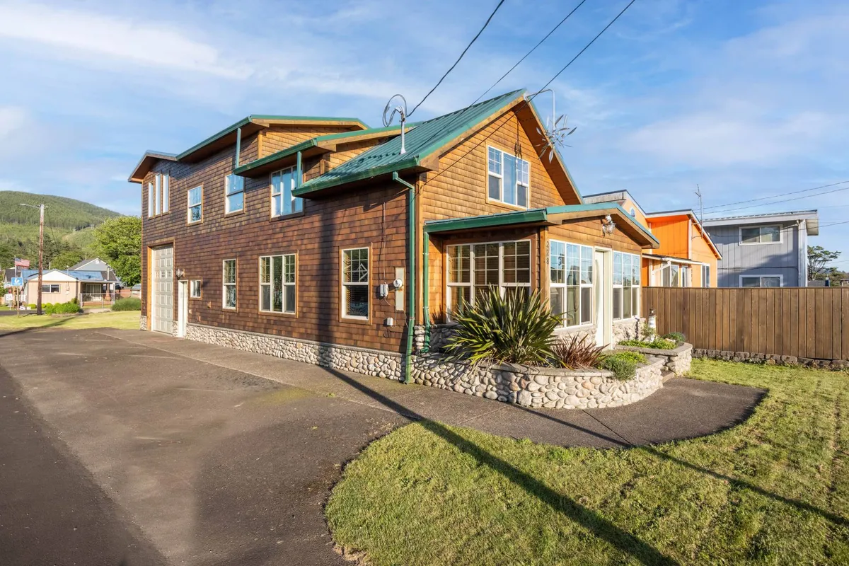 The image showcases the exterior of Cedar Lodge at Rockaway, a charming single-family house with a warm wooden facade, green roof, and beautifully landscaped garden. The inviting porch is perfect for relaxing, while the overall scene exudes a welcoming coastal retreat vibe just steps from Rockaway Beach.