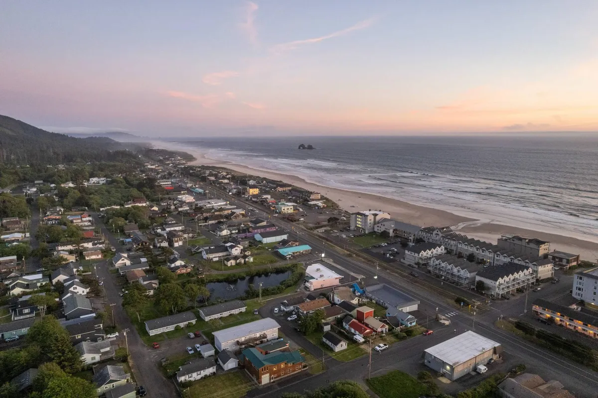 A sweeping coastal scene shows the town layout alongside the beach and ocean, under a serene sunset sky. The image captures the transition from the lively community to the tranquil shoreline, perfect for sunset walks and community gatherings.