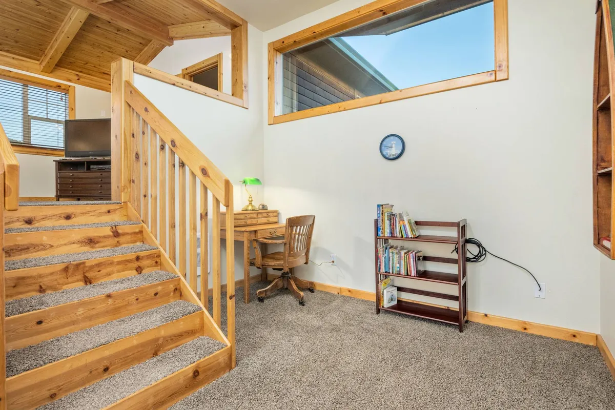 This image presents a cozy reading nook or work space, including a wooden staircase, desk with chair, bookshelf, and large window. It feels tucked away yet airy, ideal for recharging with a book or catching up on work while enjoying natural light.