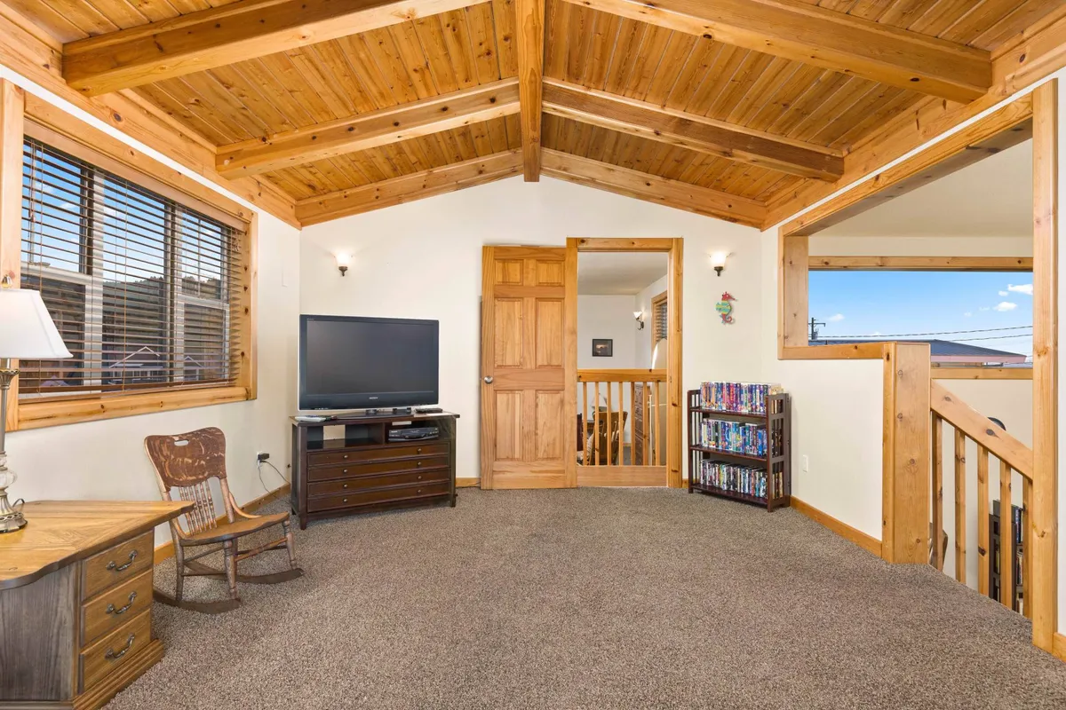 A welcoming bonus living area featuring a wooden beam ceiling, a TV, a rocking chair, and a bookshelf. The setup invites relaxation and family gatherings, evoking the nostalgia of movie nights and cozy conversations.