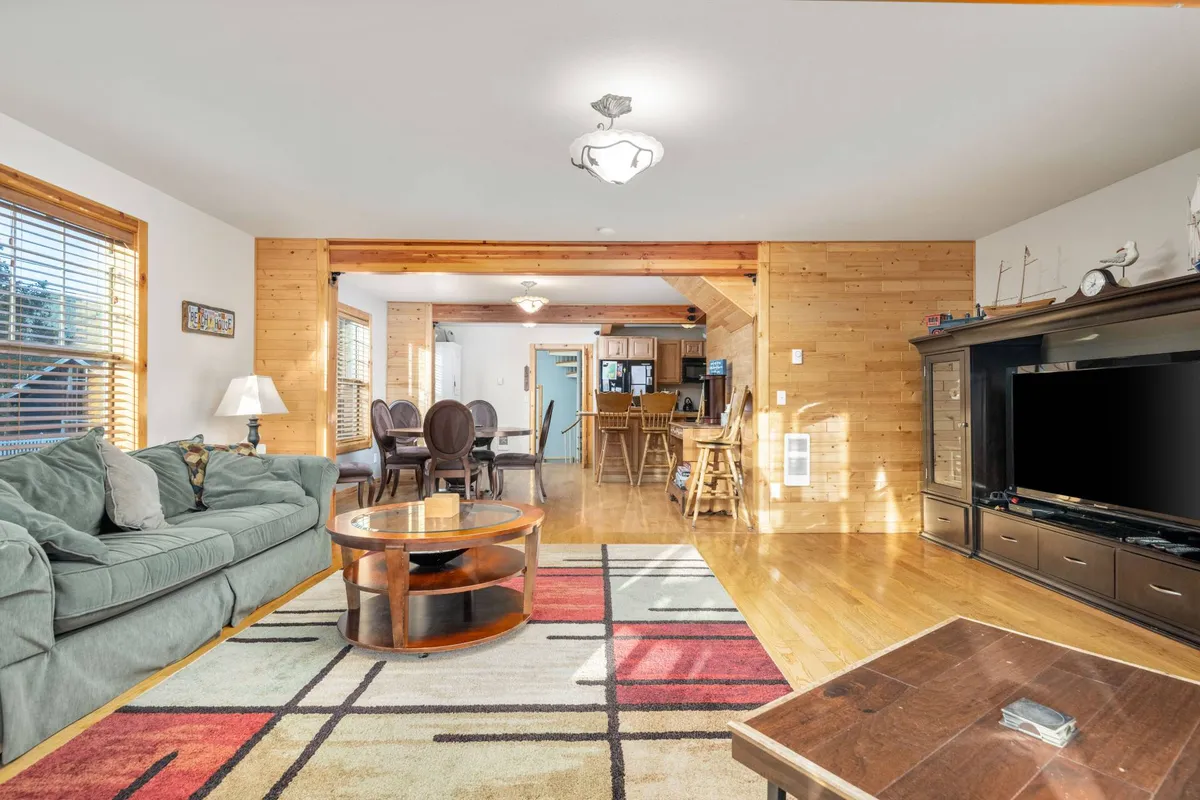 This image showcases a cozy, open-concept living area featuring a comfortable sofa, coffee table, dining area, and a glimpse of the kitchen entrance. The scene is warmly lit and arranged for gatherings, casual relaxation, or entertaining guests, with natural wood accents visible throughout.