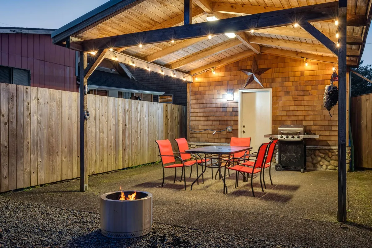 A cozy backyard scene featuring a central firepit surrounded by seating, an outdoor dining set, a grill, warm hanging lights, and rustic wooden paneling. The space is clearly fenced and designed for gatherings, with ample room for both people and pets.