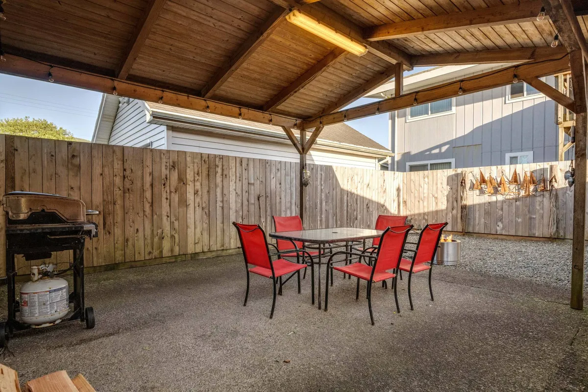 This exterior shot captures a relaxed outdoor dining space with bright red chairs and a table set beneath a sturdy, wooden-beamed structure. A grill stands ready for family barbecue nights, while the setup exudes a cheerful, communal atmosphere ideal for memorable evenings.
