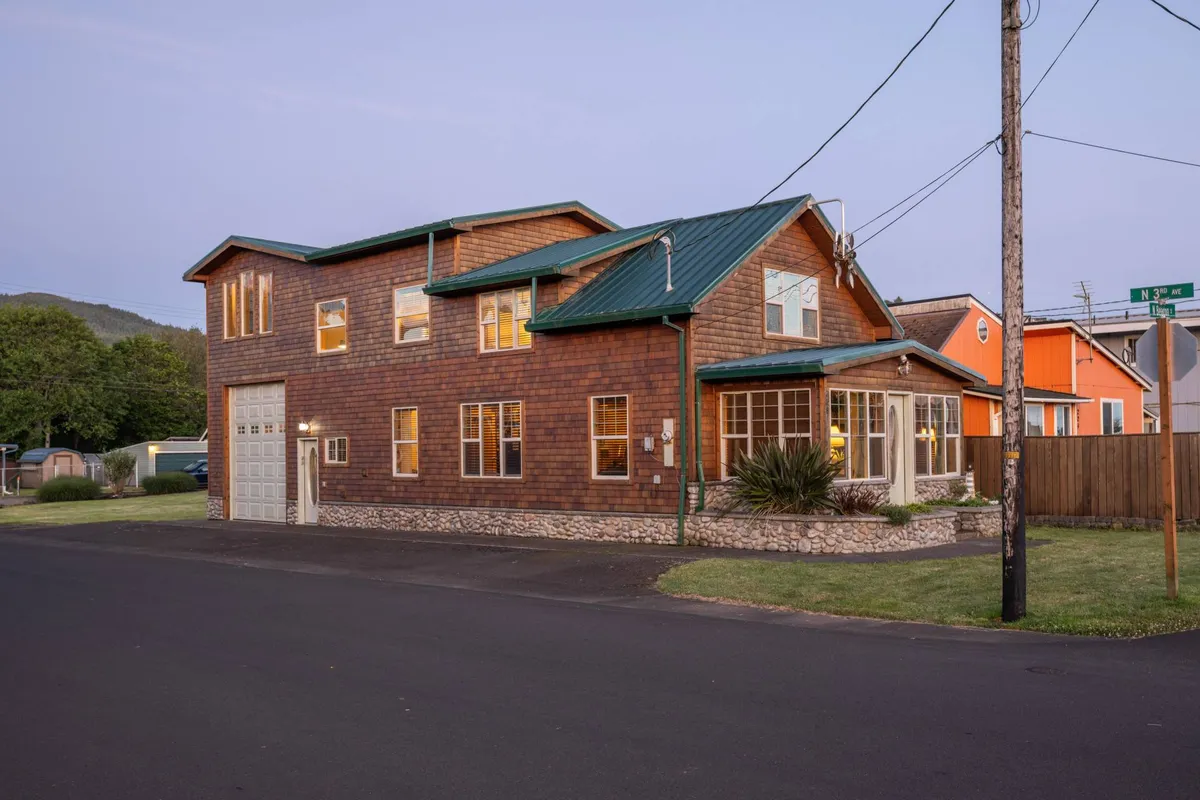 The exterior of Cedar Lodge is showcased, featuring classic wooden siding, a green metal roof, large windows, and a spacious front porch. The scene is bathed in the warm light of sunset, suggesting an inviting spot for evening relaxation after a day exploring outdoors.