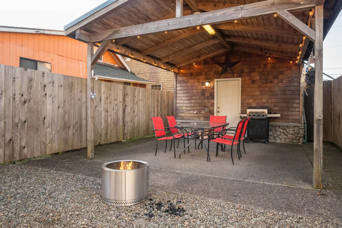 The image displays a welcoming backyard scene with a central fire pit, surrounded by comfortable seating. The area features a dining table, grill, and sturdy wooden beams supporting a covered patio, ideal for gatherings in various weather conditions. The warm glow of the fire pit and the inviting arrangement suggest a perfect setting for evening activities like roasting marshmallows and sharing stories.
