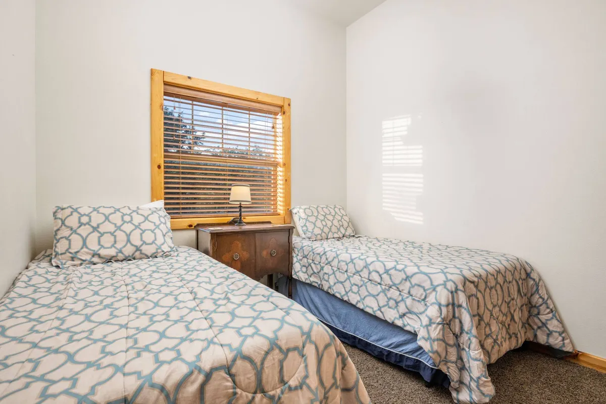 The image shows a bedroom with two twin beds situated side by side, a window with blinds letting in natural light, and a nightstand with a lamp between them. The room is softly lit, creating an inviting and serene mood—ideal for children's sleepovers or siblings sharing space.