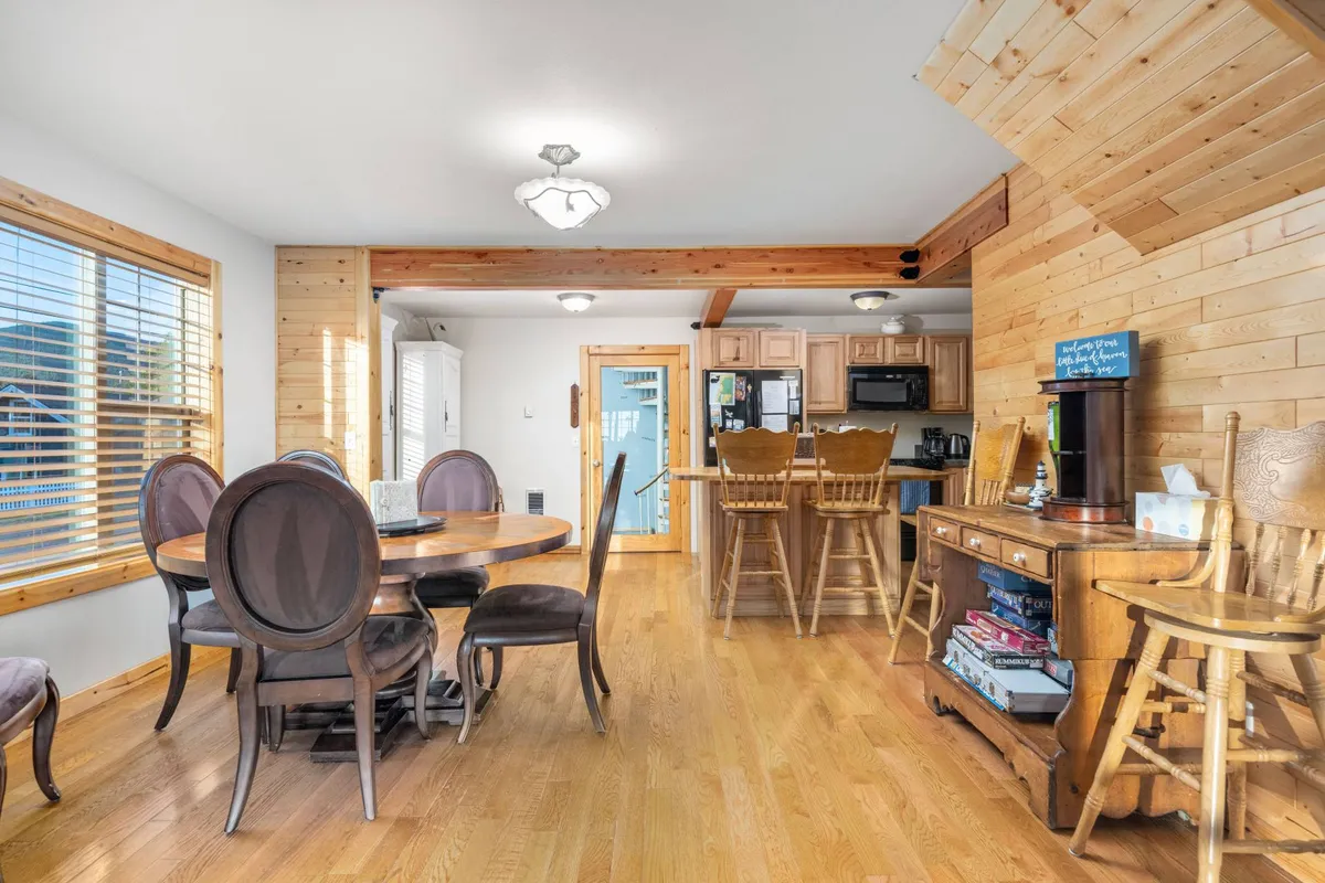 The photo features a round dining table at the center, complemented by a wooden kitchen island and cabinetry. Large windows let in abundant natural light, and stylish light fixtures add a custom touch, framing the space as both functional and welcoming for meals, conversations, or quiet moments.